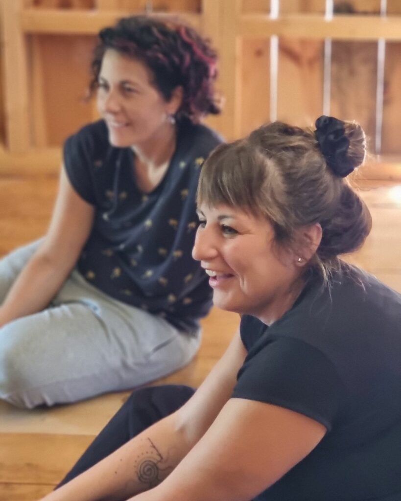 Due partecipanti del corso sedute sul pavimento di legno, sorridono mentre osservano un esercizio di lato.