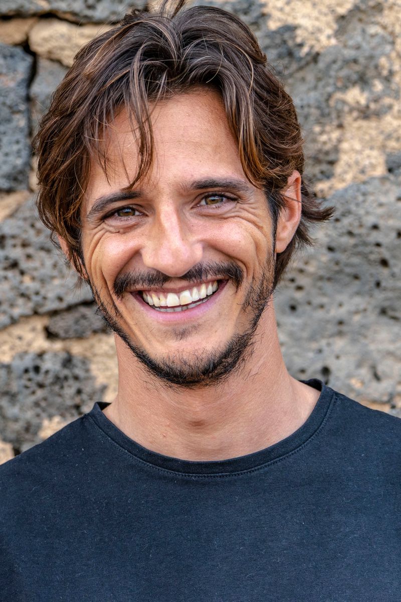 Matteo Leoni sorride verso la macchina, a mezzo busto, capelli castani mossi e barba corta, con una maglietta scura davanti a un muro di pietra vulcanica scura.