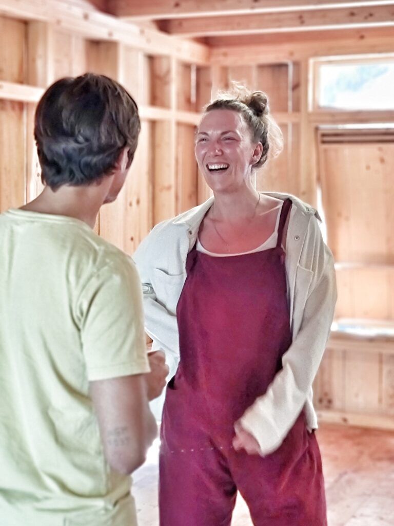 Una partecipante del corso, in salopette rosa polvere, ride di gusto davanti a un’altra persona, in una sala rivestita di legno chiaro.