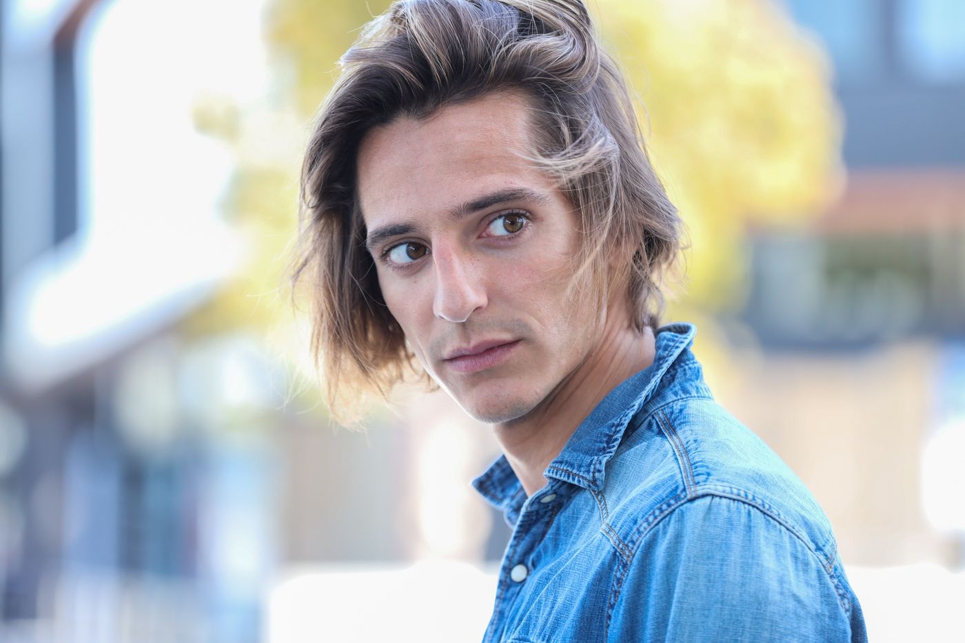 Matteo Leoni in camicia di jeans, con i capelli sul viso, si volta a guardare sopra la spalla, con una strada sfocata alle spalle.