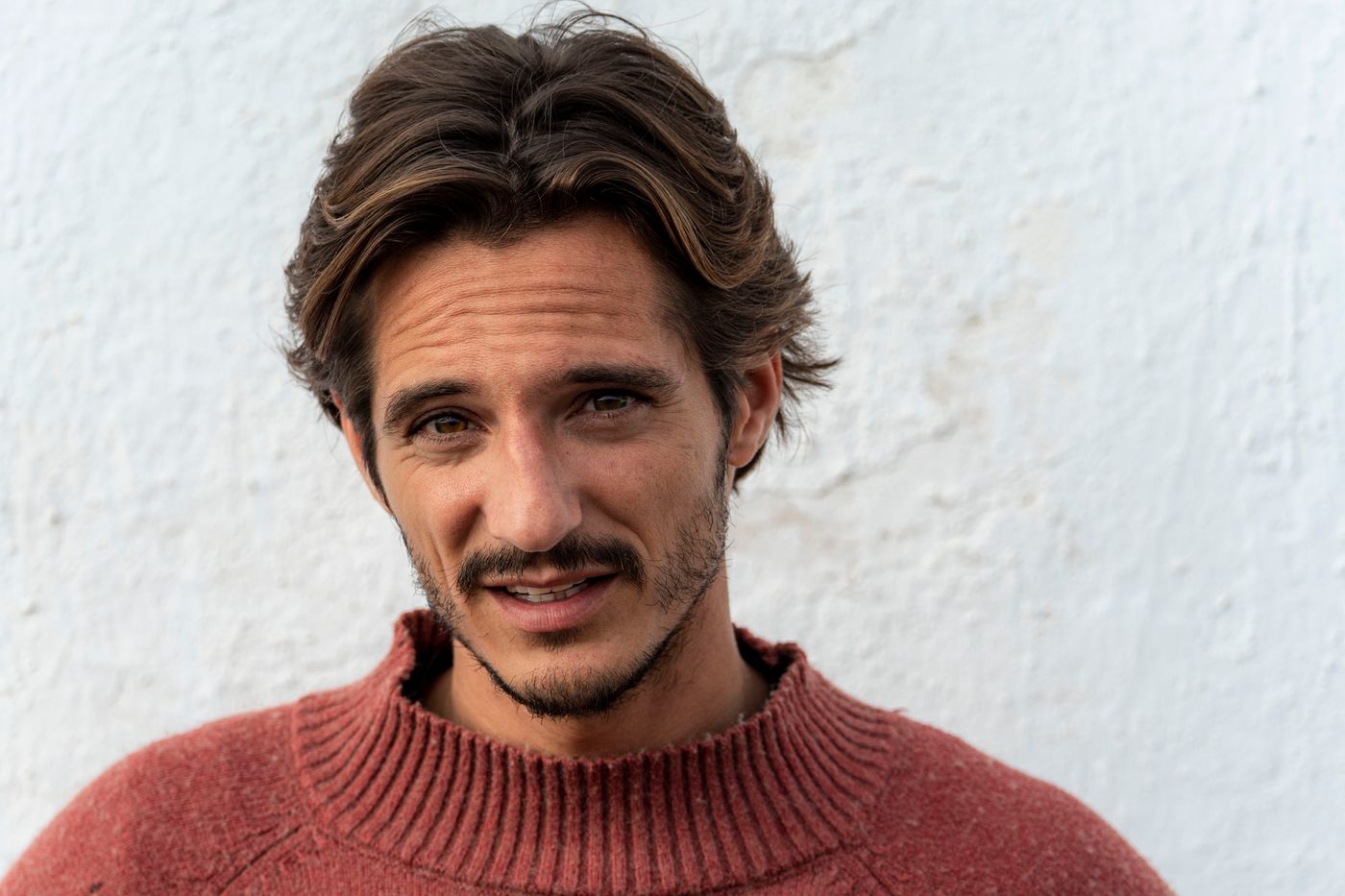 Matteo Leoni sorride a bocca socchiusa, capelli mossi sciolti, con un maglione rosso mattone davanti a un muro bianco.