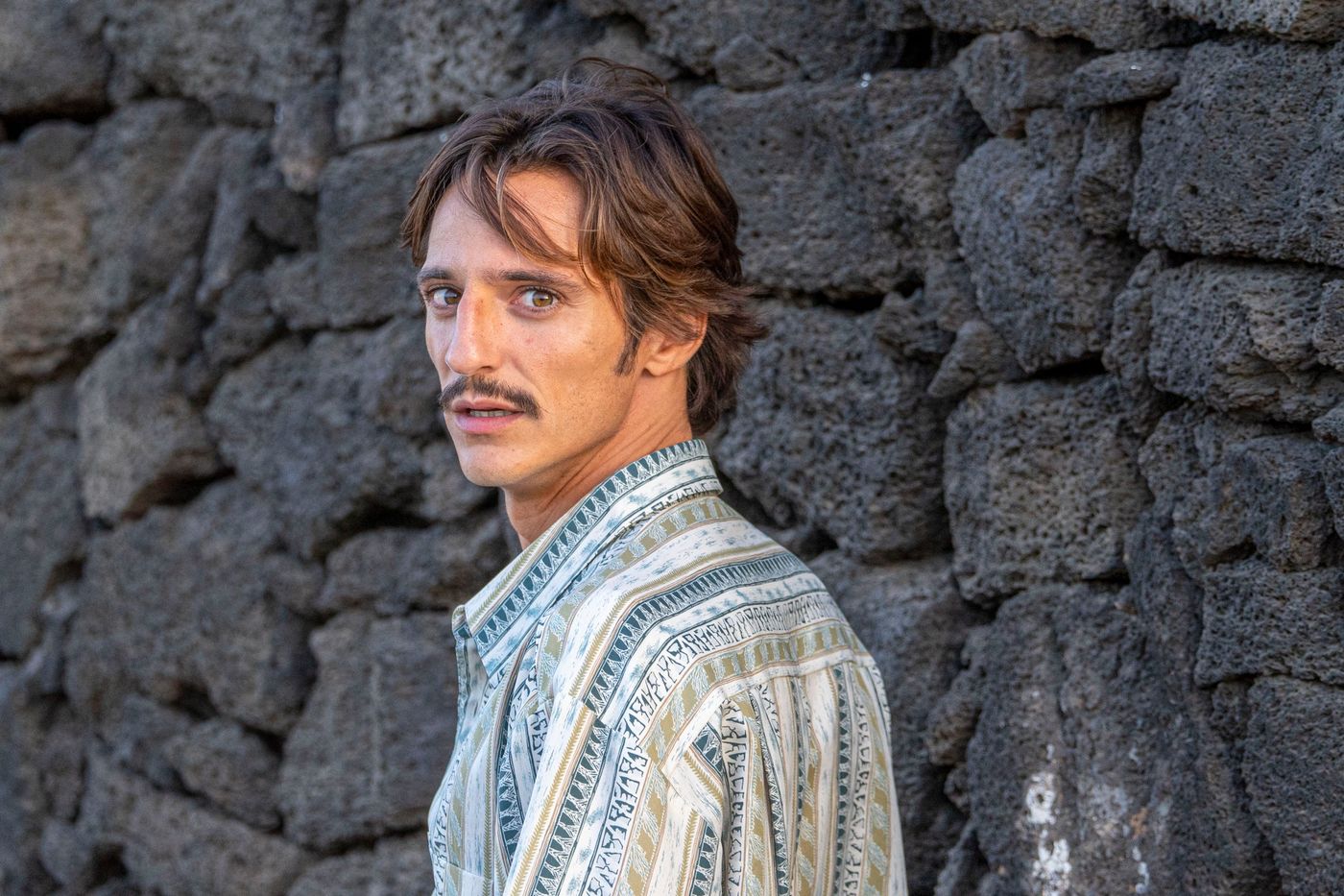 Matteo Leoni con una camicia fantasia, girato di lato e con lo sguardo oltre l’obiettivo, davanti a un muro di pietra vulcanica scura.