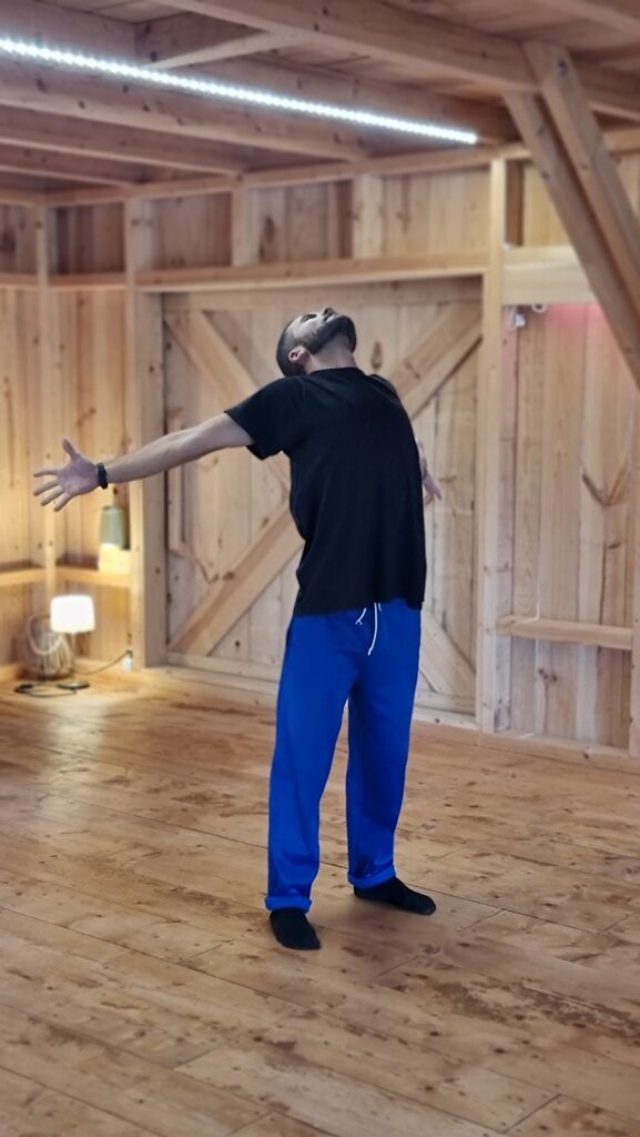 Un partecipante del corso, in calzini, in piedi al centro di una sala di legno, braccia spalancate e testa rovesciata indietro.