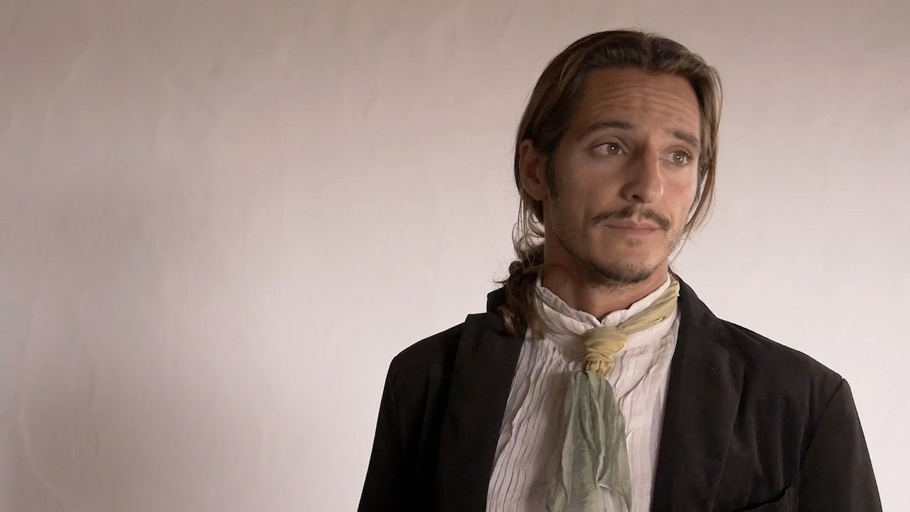 Fotogramma di scena: un uomo con giacca scura, camicia bianca e un foulard verde chiaro al collo, capelli raccolti, che guarda di lato davanti a una parete spoglia.